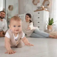 Uśmiechnięte dziecko na podłodze, ćwiczące raczkowanie. Rodzice z tyłu kibicują, zastanawiając się, czy najpierw raczkowanie czy siadanie.