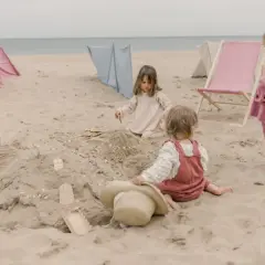 Dzieci budują zamek z piasku, ciesząc się beztroskimi zabawami na plaży. W tle widać leżaki i parawany.