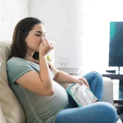 Femme enceinte s'essuyant le nez avec un mouchoir, inquiète d'un rhume enceinte danger pour bébé. Elle est assise sur un canapé, une boîte de mouchoirs à portée de main.
