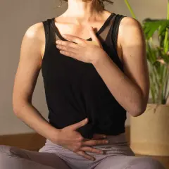 Une femme assise en tailleur, mains sur le ventre et la poitrine, pratique la respiration profonde. Une plante verte est visible en arrière-plan.