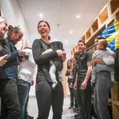 Elin Johansson, med ett leende och en bebis i bärsele, omgiven av glada människor och en svensk flagga. Kanske en paus från taekwondo-träningen?