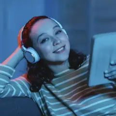 Uśmiechnięta dziewczyna w słuchawkach z tabletem w ręku, słucha muzyki, co ułatwia jej naukę angielskiego z piosenek.