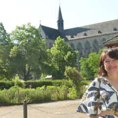 Frau lächelt vor einem Dom, umgeben von Bäumen und einem Garten. Ein Ort, der zum Verweilen und zur Entdeckung von Literatur am Dom einlädt.