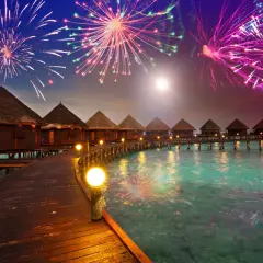 Bungalows auf Stelzen über türkisfarbenem Wasser, beleuchteter Steg, Feuerwerk am Nachthimmel. Hier ist es an Silvester warm und feierlich.