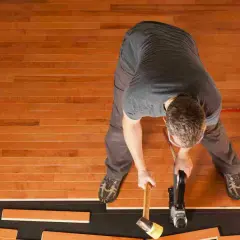 A man is installing waterproof flooring, using a nail gun and mallet to secure wooden planks.