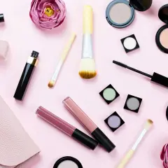 En rosa bakgrund med olika sminkprodukter som läppglans, mascara, puder och borstar. Perfekt smink i handbagage för en snabb touch-up.