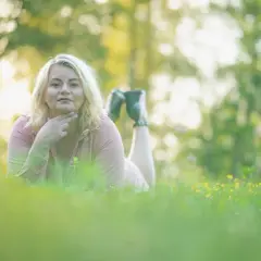 Kobieta w różowej sukience leży na trawie podczas sesji biznesowej w plenerze. W tle rozmyte drzewa i słońce.