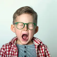 Niño con gafas verdes y camisa a cuadros, con los ojos cerrados y la boca abierta, expresando una emoción intensa, como podría ocurrir en el síndrome de Tourette en niños.