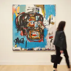 A woman walks past a vibrant, abstract painting, a striking example of Basquiat's famous paintings, featuring a bold, stylized face.