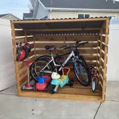 A DIY bike rack made of wood stores two adult bikes, a tricycle, and two ride-on toys. Helmets hang on a hook.