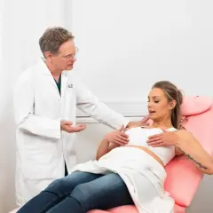 Läkare undersöker kvinnans bröst, som är klädda i en vit kompressionsbehå. Hon sitter i en rosa undersökningsstol.