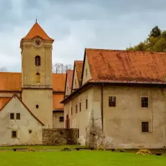 Czerwony klasztor zaprasza na zwiedzanie. Zabytkowa wieża i budynki z pomarańczowymi dachami otoczone zielenią.