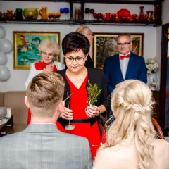 Kobieta w czerwonej sukience udziela nowoczesne błogosławieństwo parze młodej, trzymając gałązkę.
