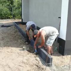 Dwóch mężczyzn układa obrzeża pod kostkę. To kluczowy etap, jak zrobić suchy beton pod kostkę, by nawierzchnia była trwała.