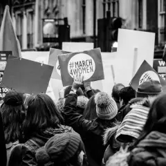 Tłum na marszu, gdzie feminizm i emancypacja idą w parze. Ludzie niosą transparenty, jeden z nich głosi "#MARCH 4WOMEN".