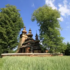 Drewniana cerkiew z kopułami na tle zieleni i błękitnego nieba, otoczona polami zbóż. Widok na Beskid Niski.