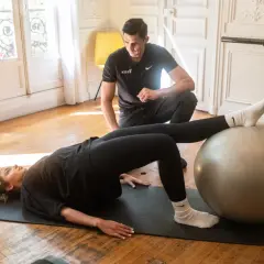 Un homme observe une femme faire un pont sur un tapis, les pieds sur un ballon de gym. Un exercice pour le bienfait d'étirement et de renforcement.