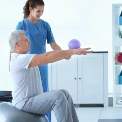 Fizjoterapeuta pomaga starszemu mężczyźnie w ćwiczeniach izometrycznych na piłce gimnastycznej.
