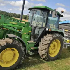 Zielony traktor John Deere 2140 z ładowaczem czołowym i przyczepą na tle łąki i lasu.