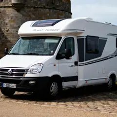 Un camping-car blanc "Dauphin Concarneau" stationné sur un sol pavé, avec un mur de pierre et la mer en arrière-plan. Un spot camping car Bretagne idéal pour explorer la côte.