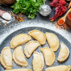 Gotowi, by nauczyć się, jak zrobić pierogi? Na talerzu surowe pierożki, obok mąka, cebula, papryczki chili i przyprawy.