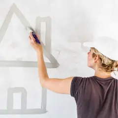 Donna con cappellino bianco dipinge una casa stilizzata su muro. Calcolare quanti litri di pittura per 50 mq.