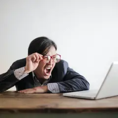 Mężczyzna w okularach krzyczy na laptopa, jakby odkrył okropne błędy w pisowni.