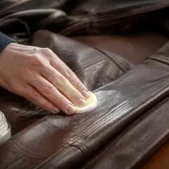 Main d'homme appliquant un produit sur une veste en cuir marron. Un pot de crème est visible. Nettoyer du cuir avec du lait est une astuce.