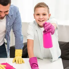 Ojciec i syn w rękawiczkach sprzątają. Chłopiec trzyma różowy spray, pokazując, jak nauczyć dziecko sprzątać.