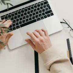 Ręce piszą na klawiaturze, obok leżą okulary i notatnik. Idealny tani laptop na studia, by zacząć naukę.