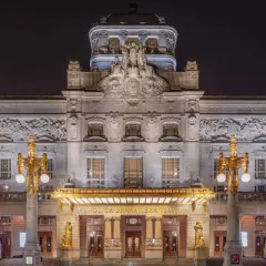 Kungliga Dramatiska Teatern i Stockholm, upplyst på natten. En storslagen byggnad med detaljerad fasad och gyllene utsmyckningar.