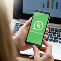 Dłoń trzyma smartfon z zielonym ekranem i logo WhatsApp Business. W tle widać laptop z wykresem.