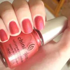 En hand med naglar målade i en glittrig, korallröd nyans. En flaska China Glaze nagellack syns i förgrunden.