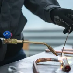 Ręce w rękawiczkach lutują miedziane rury palnikiem. Widać płomień i topiony metal, co może prowadzić do błędów przy lutowaniu miedzi.