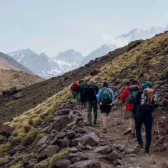 Grupa turystów wspina się kamienistym szlakiem na zboczu gór w Maroku. W tle widać ośnieżone szczyty, prawdopodobnie Jebel Toubkal.