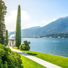 Giardino lussureggiante con gazebo bianco e cipressi slanciati, affacciato sul blu intenso del lago di Como, con montagne verdi sullo sfondo.