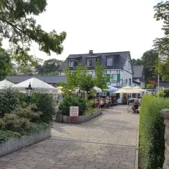 Gemütlicher Biergarten mit Sonnenschirmen und Gästen. Ein Schild weist auf den "Kleine Schweiz Velbert Rundweg" hin.