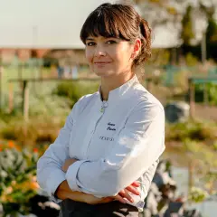 Chiara Pavan, chef con grembiule e giacca bianca, sorride con le braccia conserte in un orto.