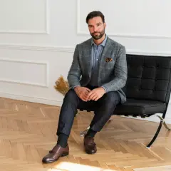 Elegancki mężczyzna w szarej marynarce i granatowych spodniach, z brązowymi butami do jeansów i marynarki, siedzi na czarnym fotelu.
