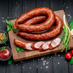Wędzona kiełbasa na desce, obok czosnek, pomidorki, musztarda i ketchup. Zastanawiasz się, ile majeranku na kilogram kiełbasy?