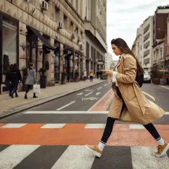Kobieta w beżowym płaszczu i żółtych sneakersach przechodzi przez pasy. Idealne buty do trencza na miejski spacer.