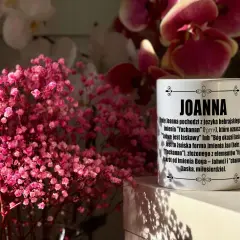 Kiedy imieniny Joanny? Na kubku z imieniem Joanna wyjaśnienie jego pochodzenia. W tle różowe kwiaty.