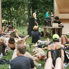 Un gruppo di persone rilassate sull'erba, ascoltando musica a un butik festival. Un DJ mixa con un laptop, circondato da altoparlanti e una struttura in legno.