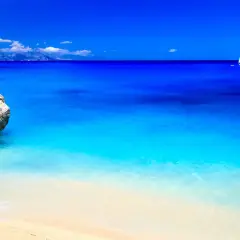 Spiaggia dorata bagnata da un mare cristallino dalle sfumature turchesi e blu intenso. In lontananza, una barca a vela solca l'orizzonte.