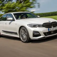 Białe BMW 320d xDrive pędzi po drodze, otoczone zielenią pól i drzew.
