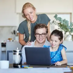 Rodzina cieszy się wspólnym czasem, tata na urlopie rodzicielskim uczy córkę korzystać z laptopa, mama wspiera ich z tyłu.