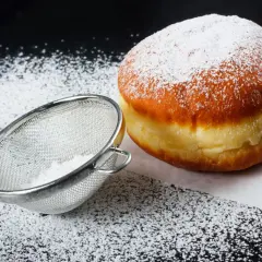 Puszysty pączek posypany cukrem pudrem, obok sitko z cukrem. Idealne na tłusty czwartek, pyszne pączki!