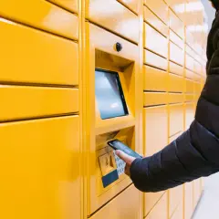 Osoba w czarnej kurtce zbliża telefon do paczkomatu. Zastanawia się, czy jak nie odbiore paczki to dostane zwrot pieniedzy.
