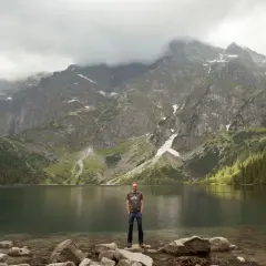 Mężczyzna stoi na kamieniach nad brzegiem jeziora Morskie Oko. W tle majestatyczne góry Tatr.