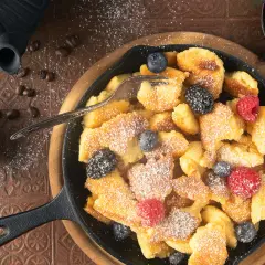 Ein herzhafter gerupfter Pfannkuchen, bestreut mit Puderzucker und garniert mit frischen Beeren, serviert in einer Gusseisenpfanne.